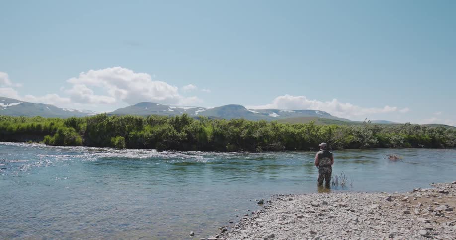 Fisherman in a clear river surrounded by vegetation and mountains in the background