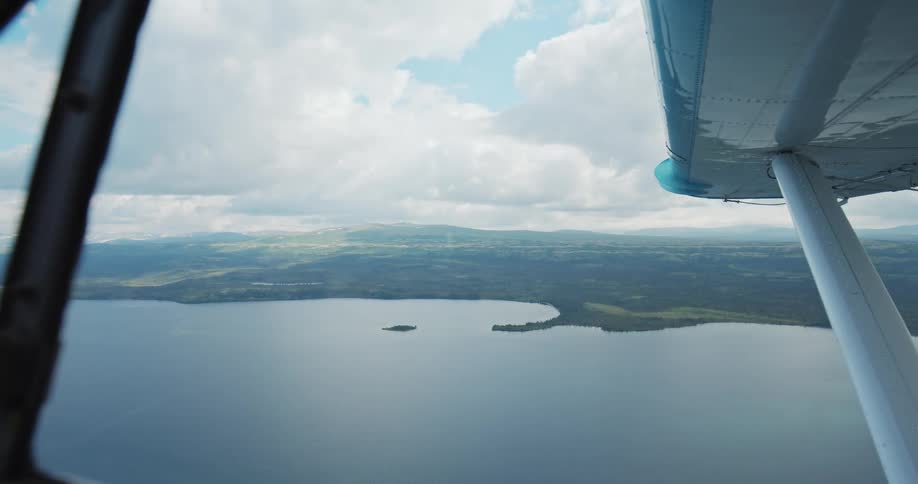 The video is an aerial view filmed from the window of a small plane showing a large lake surrounded by rolling forested hills and distant mountainsthe planes light blue and white wing and struts frame the right side of the shot while the water fea