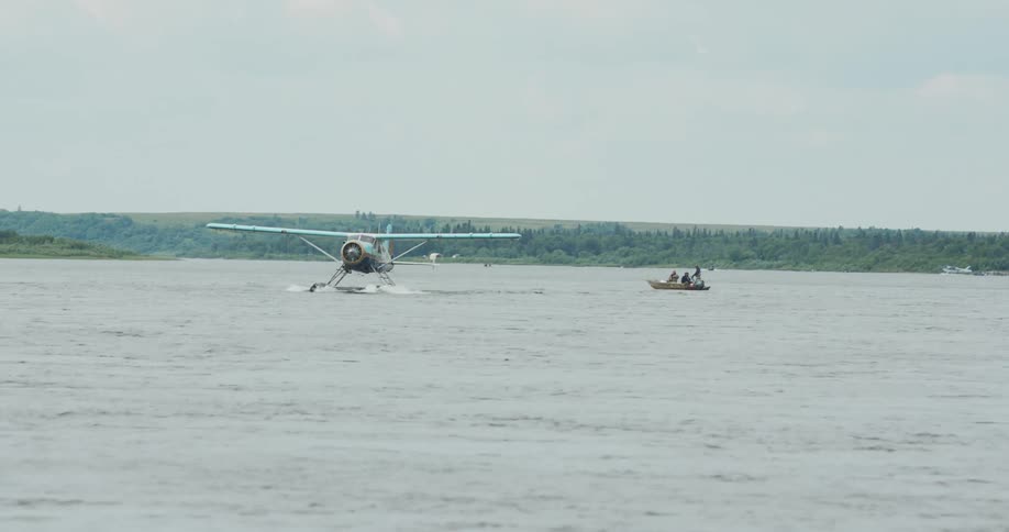 The video shows a lowangle shot of a singleengine floatplane on a wide body of water possibly a large river or lakethe plane is facing the camera and appears to be moving slowly or taxiing near a small boat carrying a few people with a wooded shore