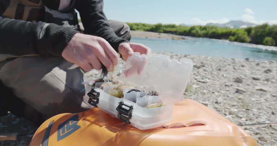 The video is a closeup shot of a person dressed in waders kneeling on a rocky riverbank and sorting through a clear plastic tackle box filled with various fishing flies and luresthe background features a beautiful view of a clear bluegreen river f