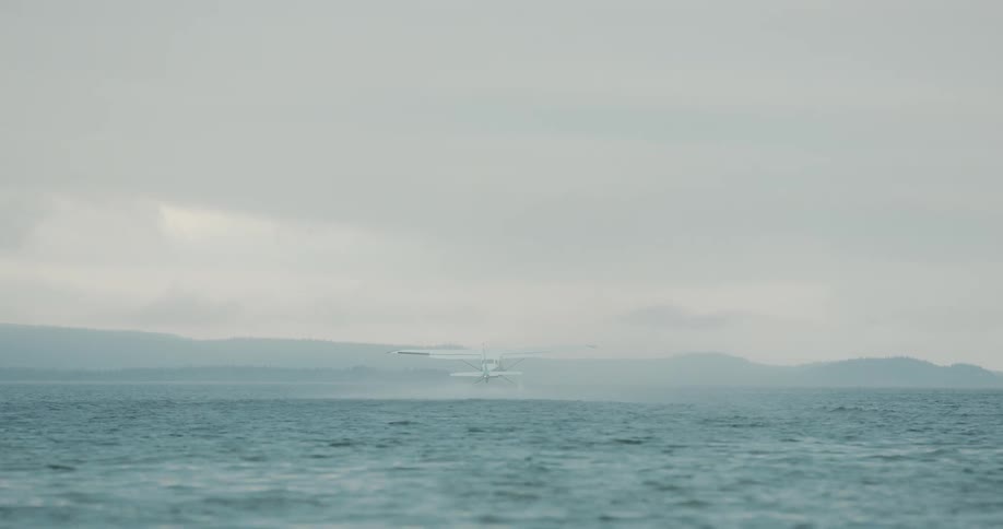 The video captures a rear view of a white and blue floatplane taking off from a large body of water like a lake or bay under an overcast skythe aircraft accelerates creating a large spray of water behind its floats before becoming airborne and flyin