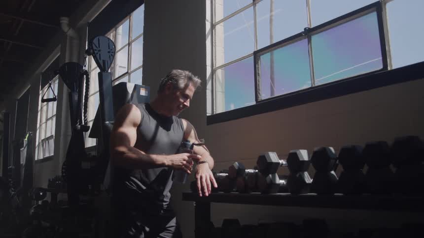 A middle aged man holding a water bottle while standing by the window in a gym after a fitness session