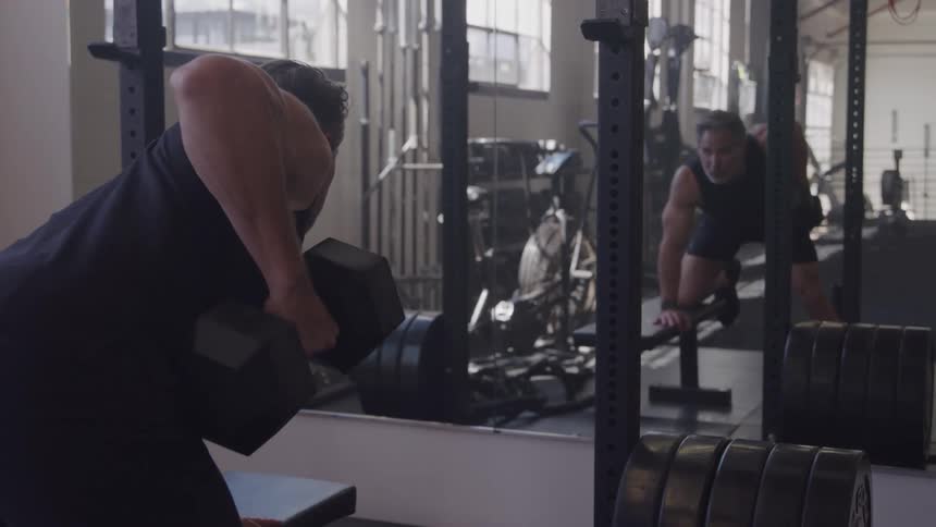 The video shows a man in a black tank top performing a dumbbell row exercise in a gym with his body bent over and his left arm lifting the weighthis reflection is visible in a large mirror opposite him which also reflects various weights and gym equi