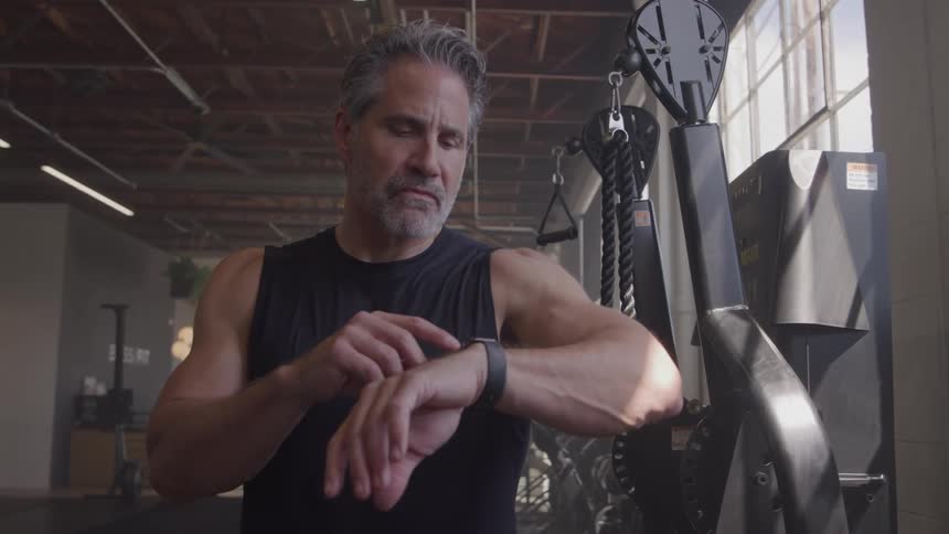 A middle age man stands next to a piece of gym equipment and looks down at the wristworn device on his left arm in an indoor exercise facility