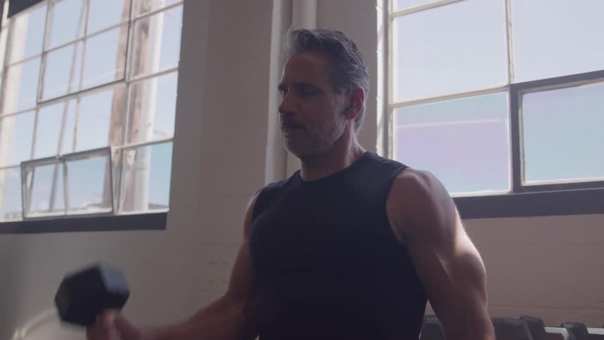 A middleaged man with massive muscles lifts a dumbbell in the gym at midday focused and intense earbuds in as he powers through his workout