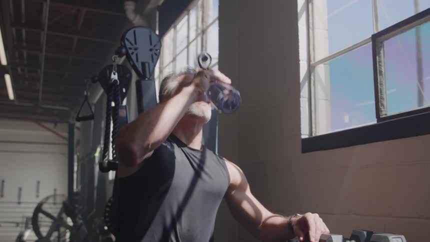 The man is shown standing next to a rack of dumbbells lifting a clear water bottle to his mouth to take a drink sunlight streams in through large windows behind him illuminating the interior of the gym