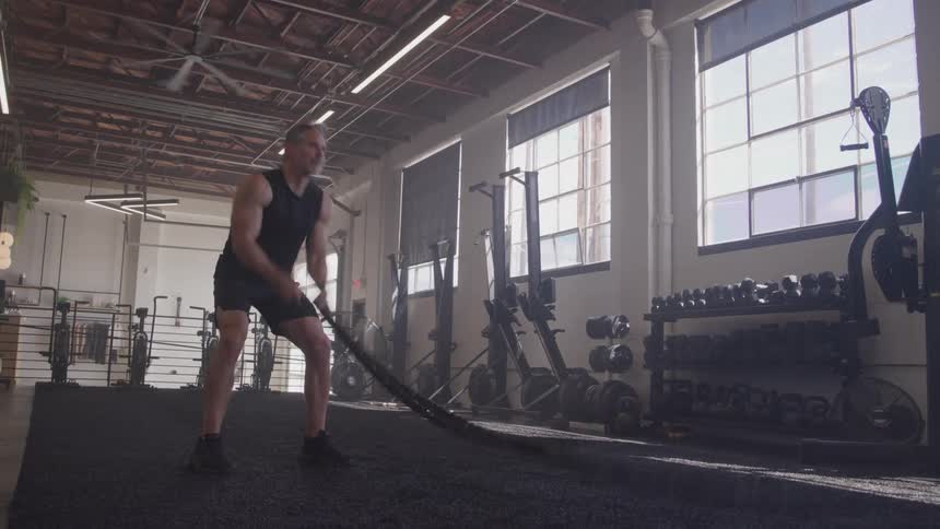 A fit middle aged man with graying hair vigorously exercises with battle ropes in a spacious well equipped gym