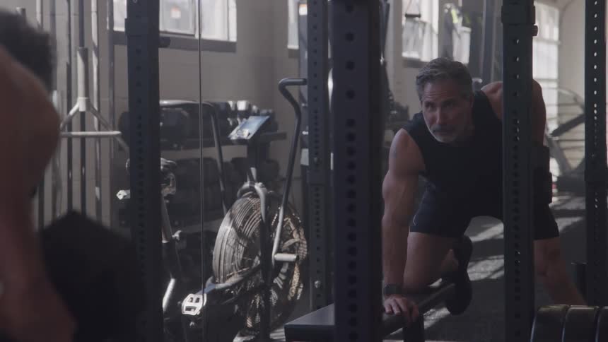 Man leaning on a gym bench while training with dumbbells in front of the mirror