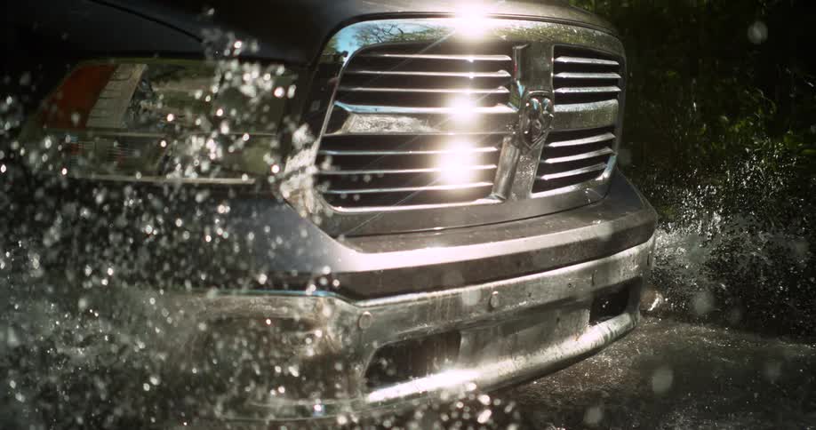 A closeup of a vehicles front bumper and headlight plows through muddy water throwing sparkling droplets into the air