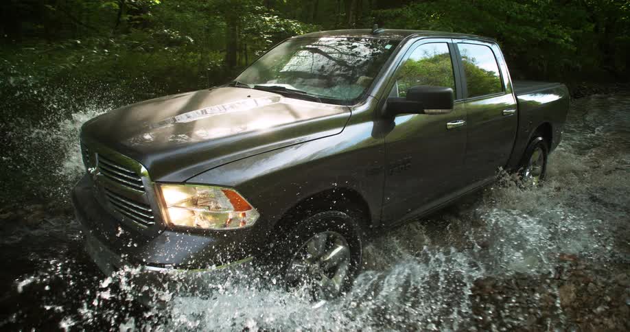 A dark gray pickup truck moves slowly through a shallow stream or ford causing a large spray of water around its wheels and chassis framed by a dense forest in the background