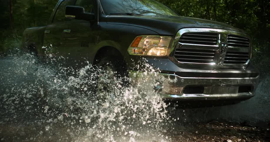 A dark gray pickup truck moves slowly through a shallow stream or ford causing a large spray of water around its wheels and chassis framed by a dense forest in the background