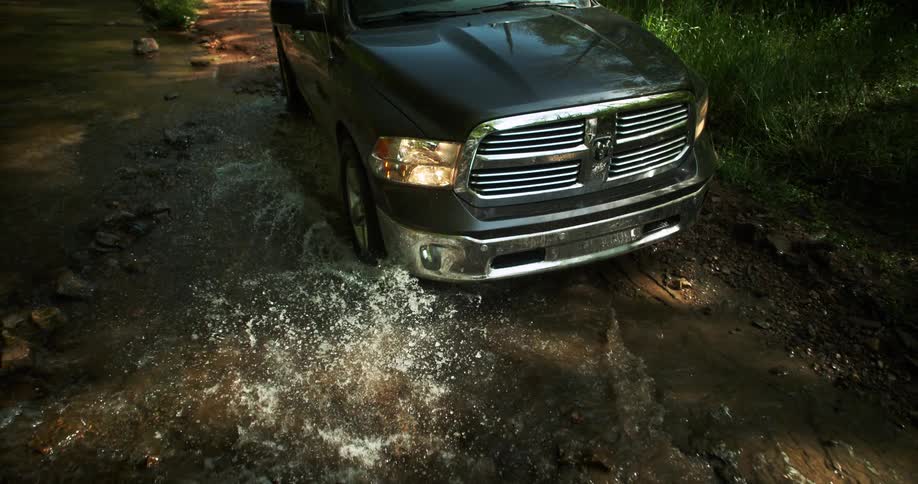 The front of a dark gray pickup truck moves slowly through a shallow puddle on a dirt road in a wooded area causing water to splash over the ground and front bumper