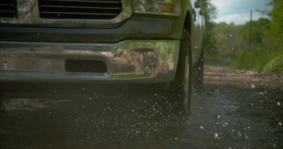 Lowangle closeup of a pickups chrome bumper and front wheel rolling along a wet muddy gravel road with puddles