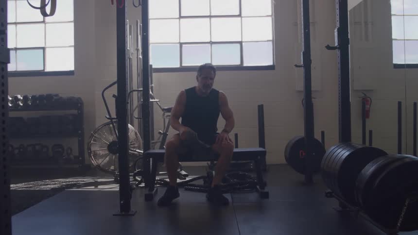 The man walks toward the center of the gym and sits down on a flat weight bench he then uses a towel to wipe the sweat from his face and neck