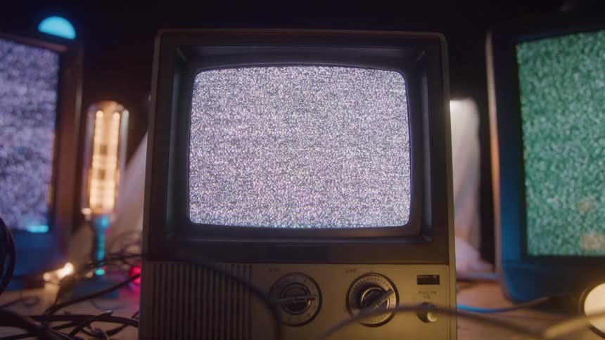 A vintage small crt television sits in the center foreground its screen filled with static or snow it is surrounded by tangled cables glowing vacuum tubes or long bulbs and other screens also displaying static