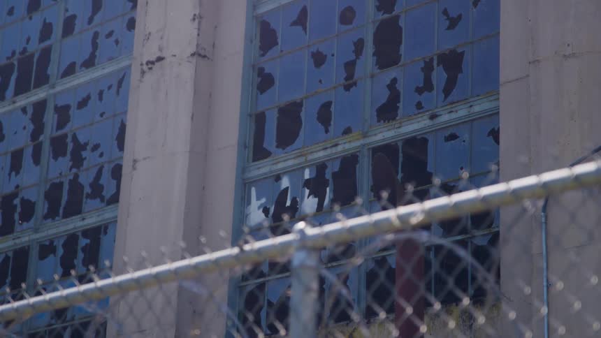 The clip shows the exterior of a building with large gridstyle windows that have numerous broken panes in the foreground a chainlink fence with strands of barbed wire runs horizontally across the bottom of the frame