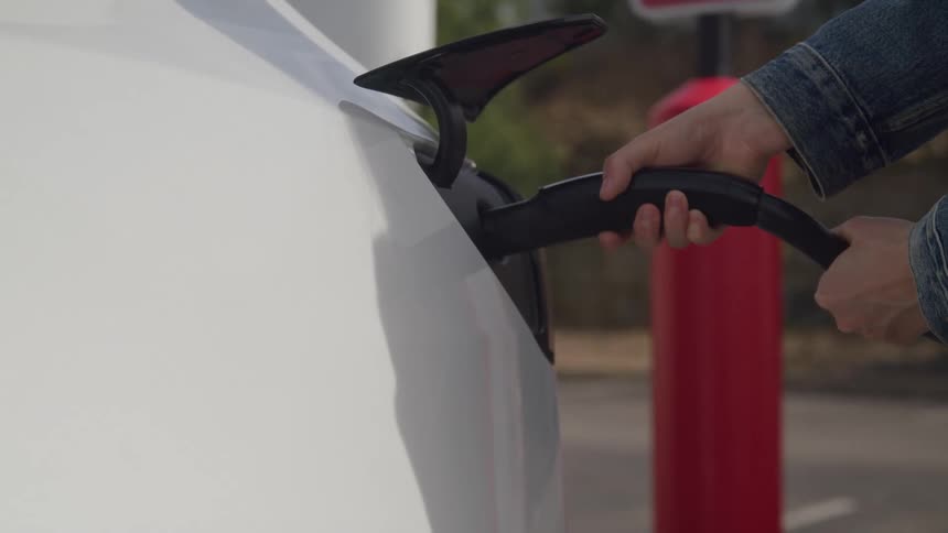 A person plugs a charger into an electric car at a charging station during midday connecting the cable to the vehicle as sunlight reflects off the polished surface