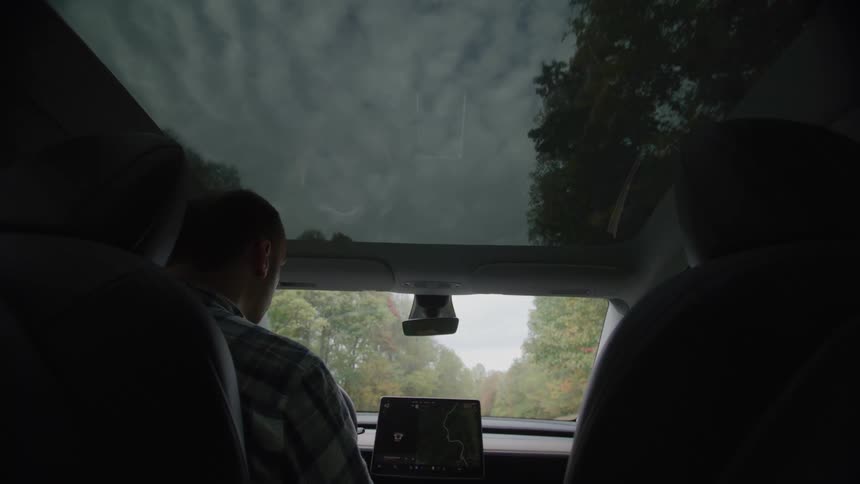 Backseat view of a car with a panoramic glass roof as a driver in a plaid shirt heads down a treelined road the center screen showing a map under an overcast sky
