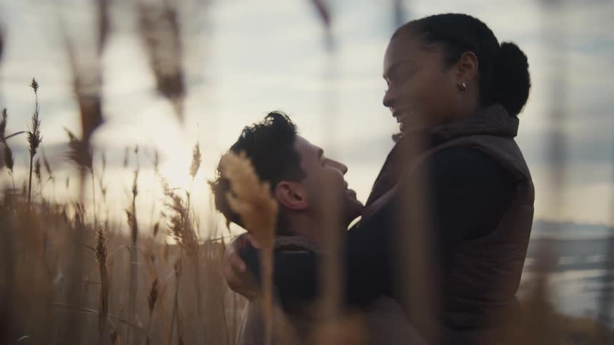 A young couple stands on a grassy shoreline at midday with the man carrying his girlfriend and spinning her around