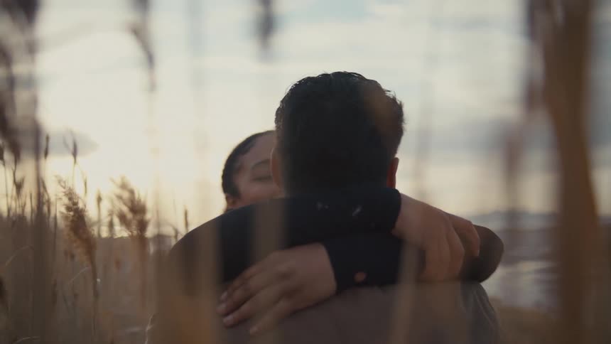 A young couple embraces in tall grasses at sunset the woman smiling playfully as the man lifts her in a warm backlit hug