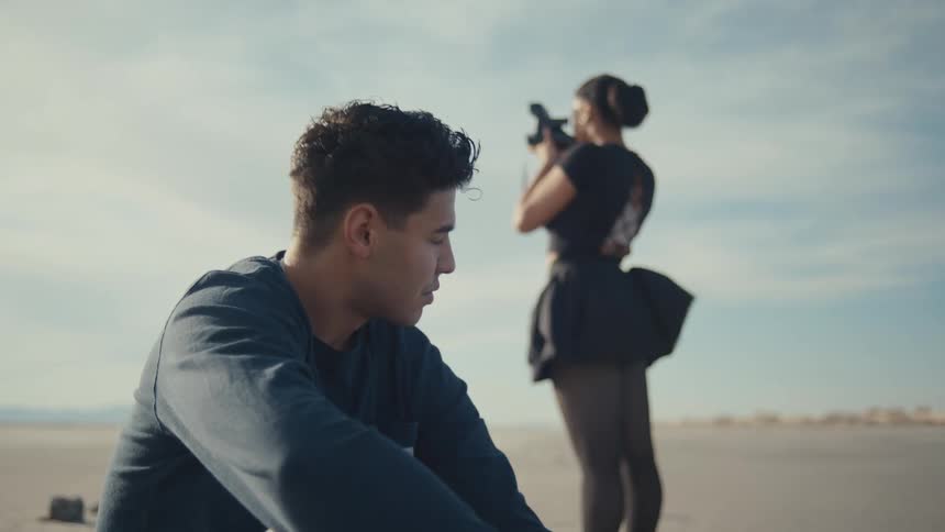 While a man rests on a desolate landscape a woman dressed in ballet attire films him