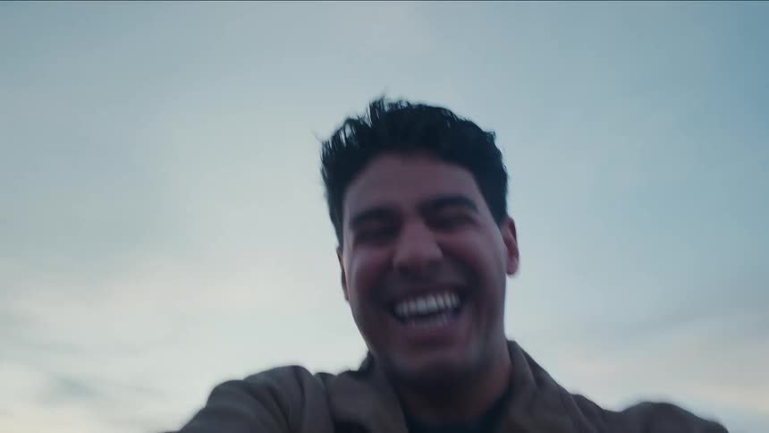 A closeup of a man with short dark hair in a brown jacket grins broadly seeming to be running in a circle seen from the arm up against a pale blue sky slight motion blur emphasizing his joyful excitement