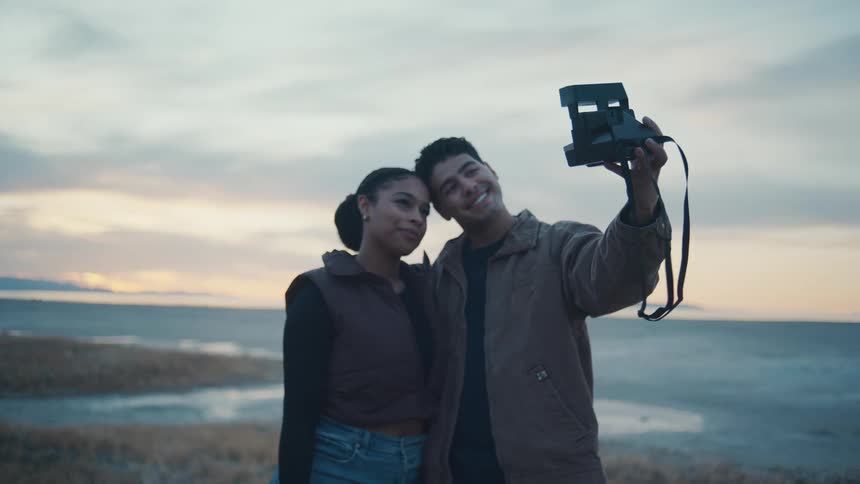 A couple in warm jackets stands by a calm shoreline at sunset leaning into each other as the man smiles and holds up a vintage instant camera for a selfie