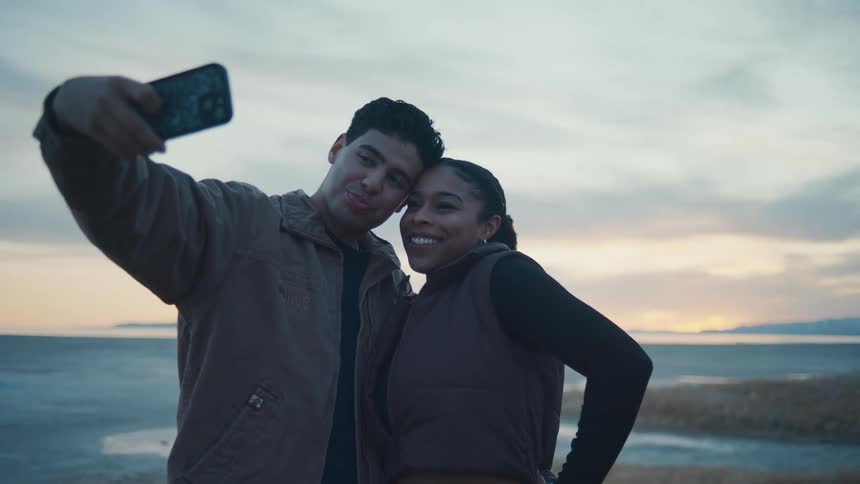 A young man and a young woman stand outdoors near a body of water hold a mobile phone up and pose for a picture as the woman gives a kiss on the males cheek