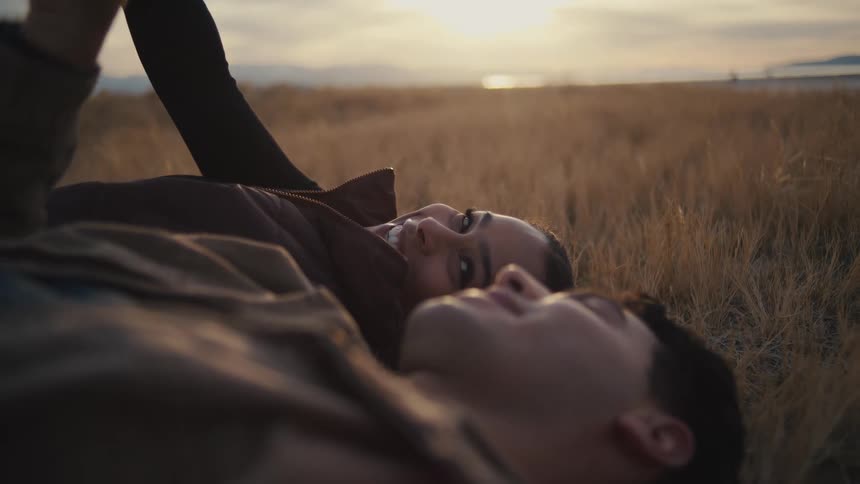 A young couple lies on a grassland at midday looking at the sky while talking and laughing together