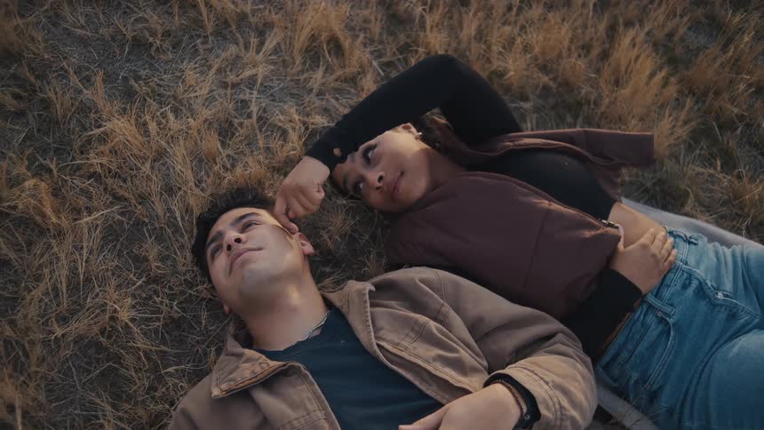 A couple lies on a grassland turned brown from fall at midday both wearing jackets as the young woman rubs her finger on the mans face