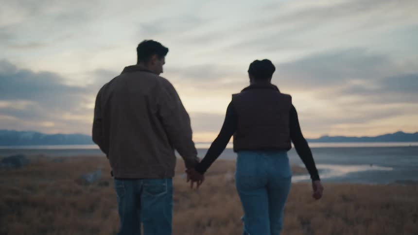A man in a brown jacket and a woman in a dark vest and blue jeans stand sidebyside holding hands on a dry grassy shore overlooking a wide lake and distant mountains under a cloudy twilight sky before they turn to face each other