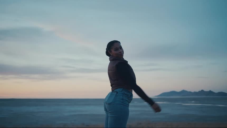 A smiling young woman wearing a dark vest and blue jeans stands outdoors raising her arms triumphantly and turning to look over her shoulder before walking toward the camera against a vast flat landscape and a dusk sky with distant mountains