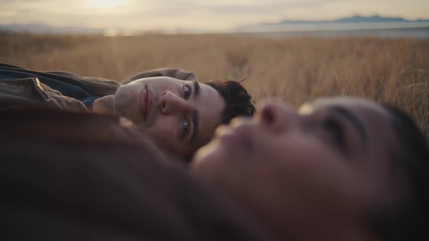 A man lies next to a woman in a field of dry grass at sunset