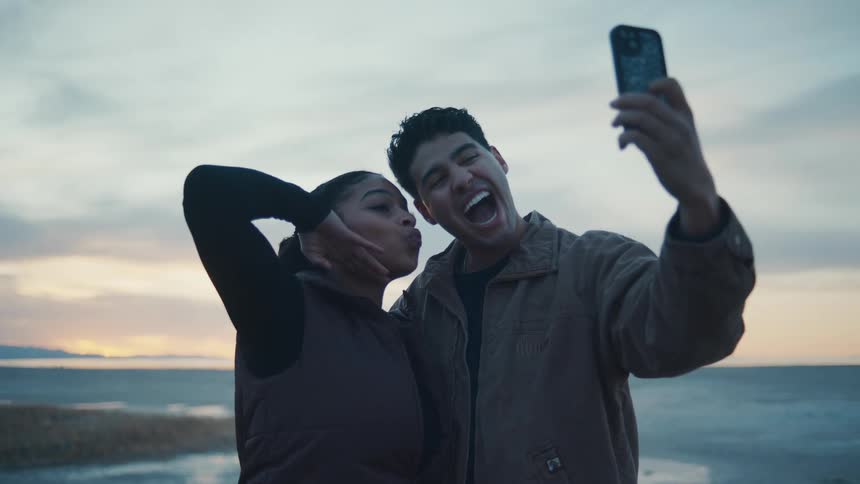 A young man and woman take a selfie by the shore at dusk both acting silly making faces