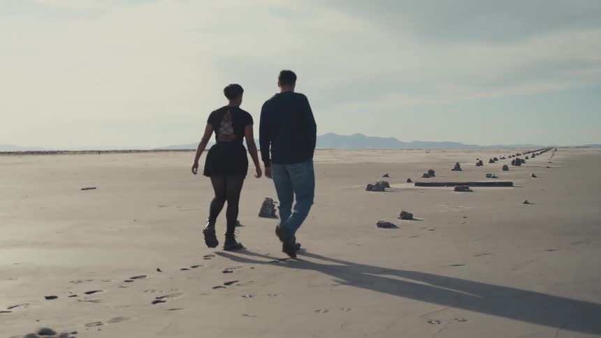 Couple walks on sandy terrain toward distant mountains under bright sky