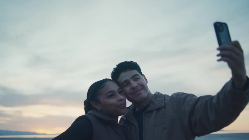Young man and young woman kissing outdoors while they take pictures on a smart phone during dusk or dawn
