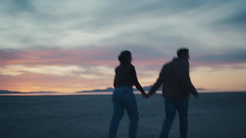 Couple runs across the beach holding hands while sunlight brightens on the red horizon