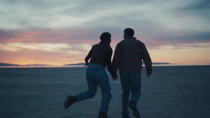 A young couple seen from behind wearing jeans and jackets walks handinhand across a vast flat dry landscape and leaps into the air together in silhouette against a dramatic orange and pink sunset sky