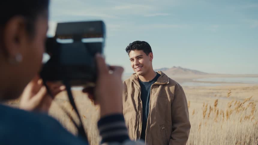 Young man posing and making goofy face expressions while a young woman takes pictures of him with a camera outdoors during day time