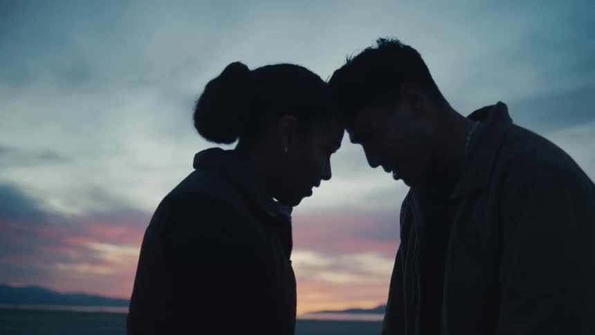 A young couple stands outdoors at sunset both wearing jackets as they bump their heads together with smiles on their faces