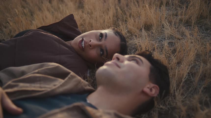 A woman smiles warmly while lying in dry grass facing a man in the foreground both in brown jackets sharing a relaxed intimate moment at dusk