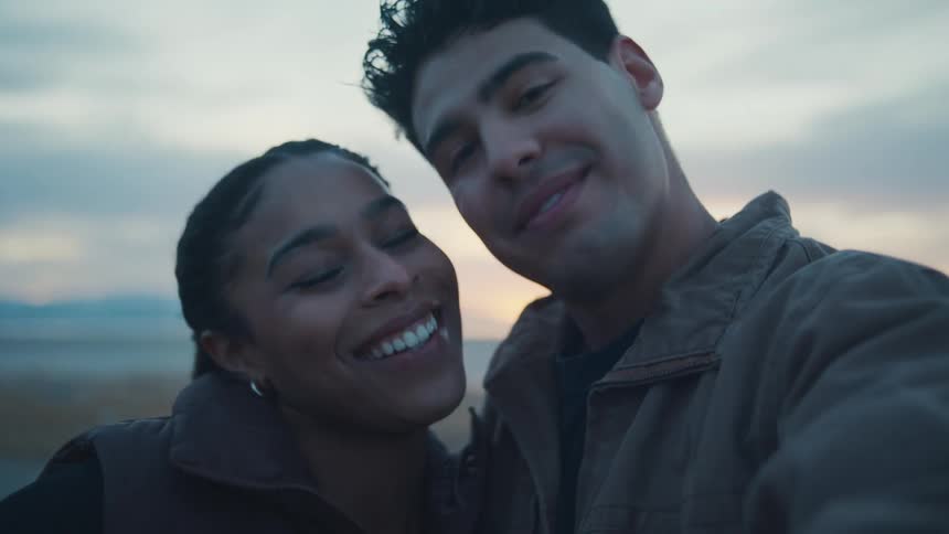 A young couple smiling pouting for a kiss and kissing on the cheek while holding the camera up for a selfie against a pale outdoor sky