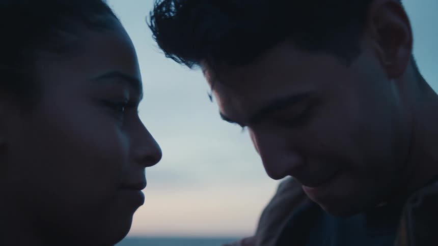 A young man and woman stand close together in a dark intimate profile view touching foreheads and smiling softly at each other with the distant horizon and pale sky providing a subtle muted background light