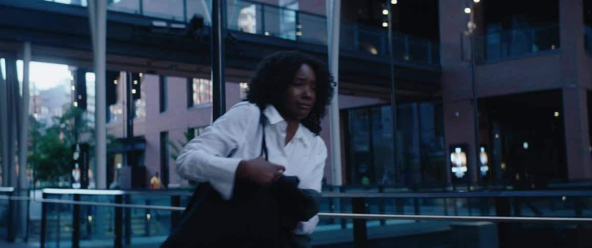 A young woman with curly dark hair wearing a white business shirt and carrying a black bag stands up as she walks quickly across a pedestrian bridge or elevated walkway in a modern dark city interior or atrium with illuminated windows