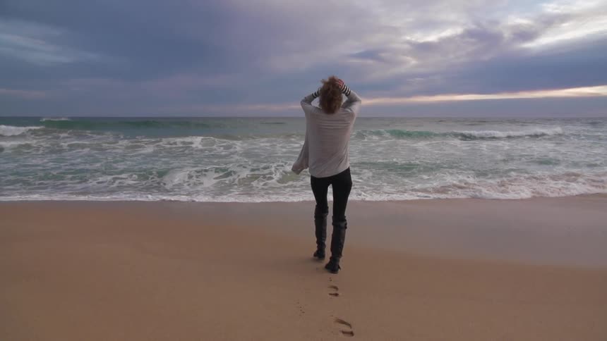 A young woman walks towards the water as large waves crash ashore and storm clouds loom in the distance