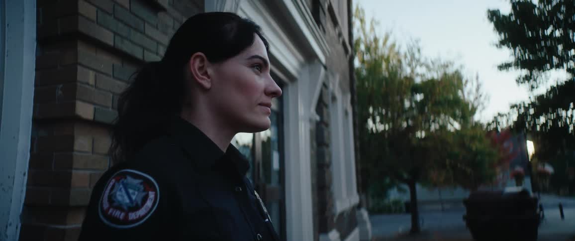 Uniformed woman with a fire department patch observing the street beside a building