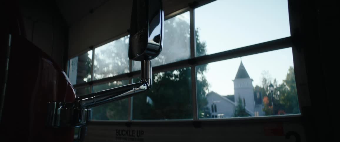 The shot looks outward from inside a garage with a chrome fixture hanging down in the foreground and a buckle up sticker visible below outside the multipaned window a church with a tall steeple is visible across the street amidst green trees