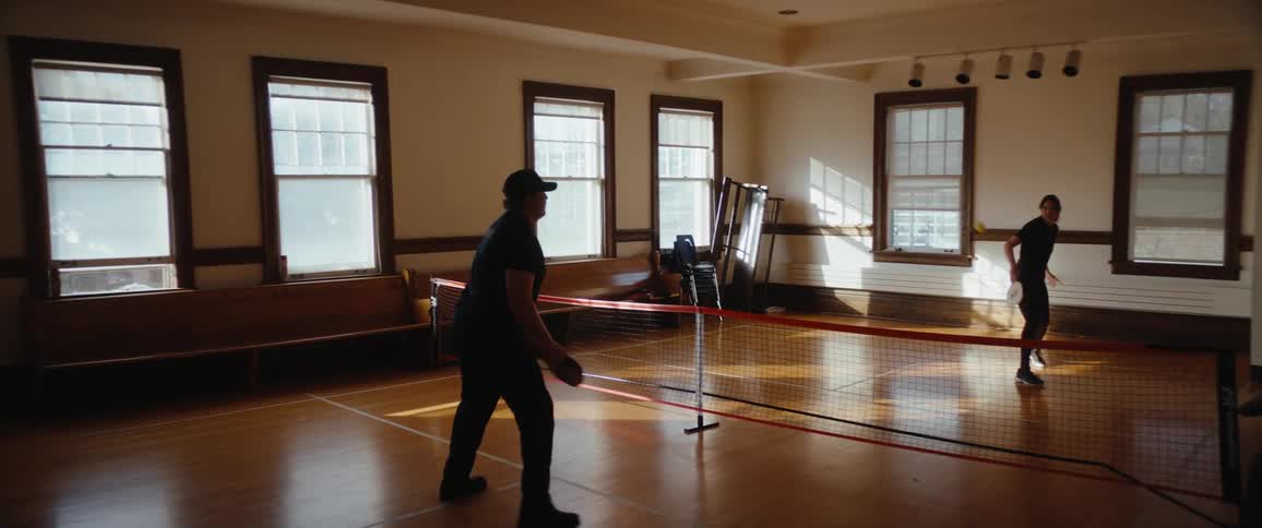 Two people are playing an indoor racket sport likely pickleball on a polished wooden floor court with a low net the room is brightly lit by sunlight streaming in through a row of tall windows along the back wall