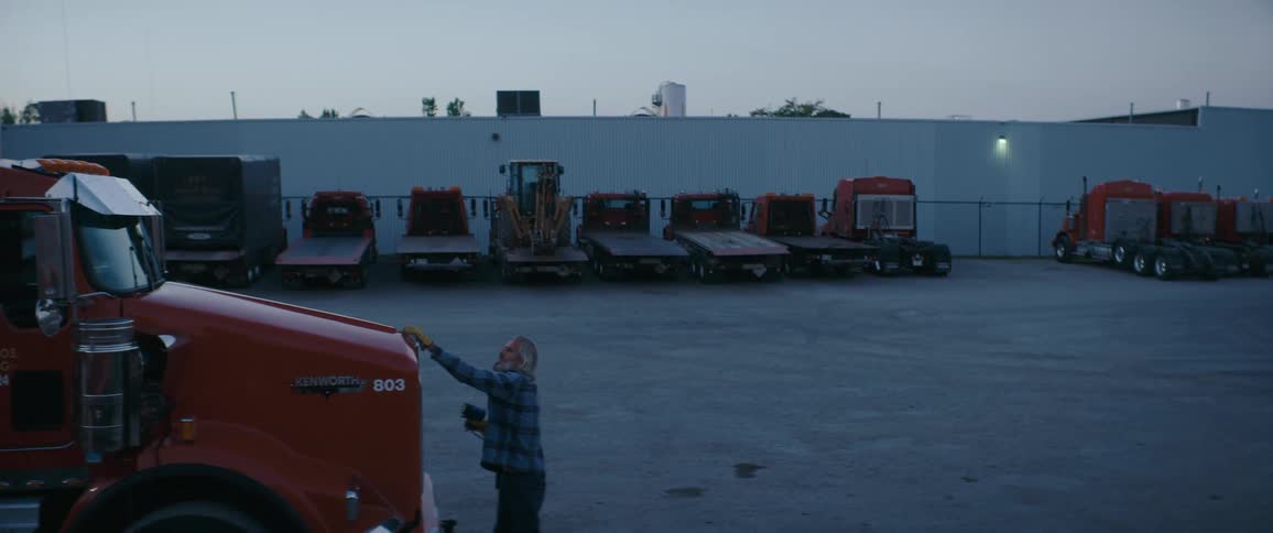 An elderly man stands in a trucking yard and lifts the hood of a semitruck in the foreground while a line of cargo trucks and trailers are parked against a white industrial building in the background