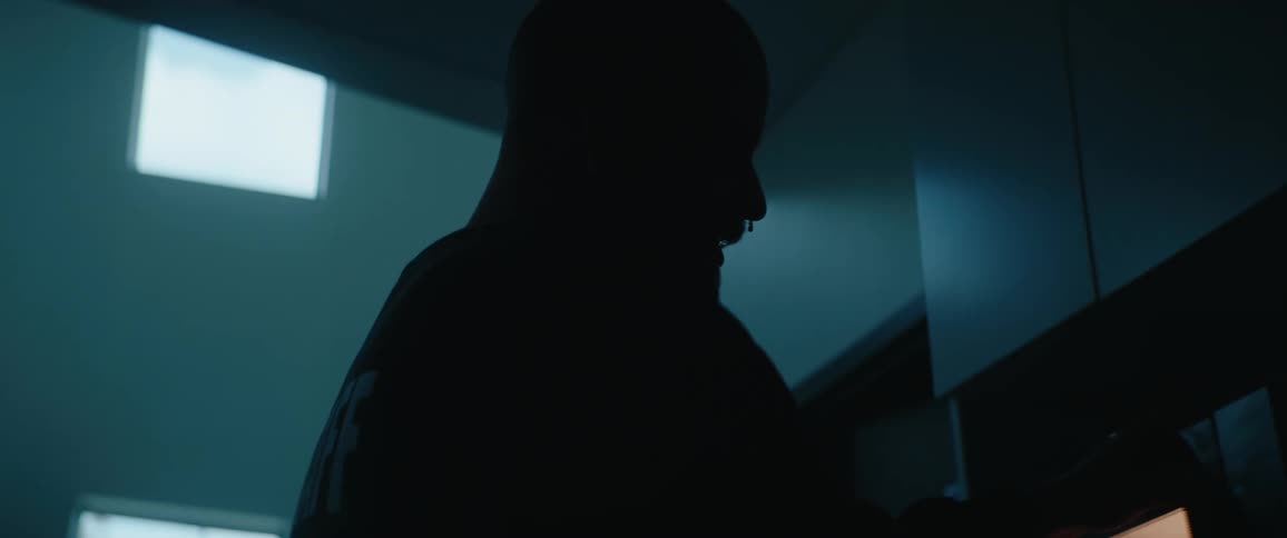 A man is seen in silhouette from the back and side standing in a dimly lit room bright possibly fluorescent light comes from windows or a fixture behind him creating a strong rim light effect